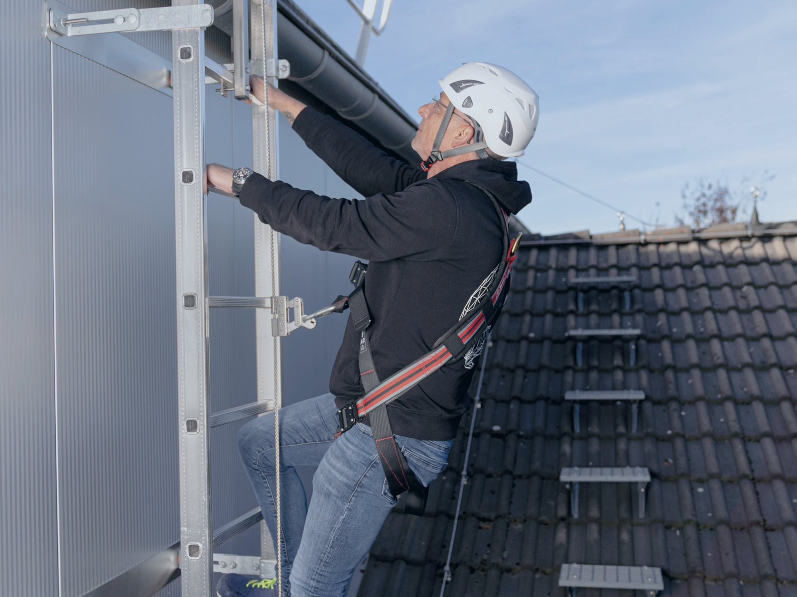 Person med faldsikringsudstyr klatrer på fast stige med vertikalt livlinesystem og glider, som sikrer kontinuerlig faldsikring under op- og nedstigning på tag.