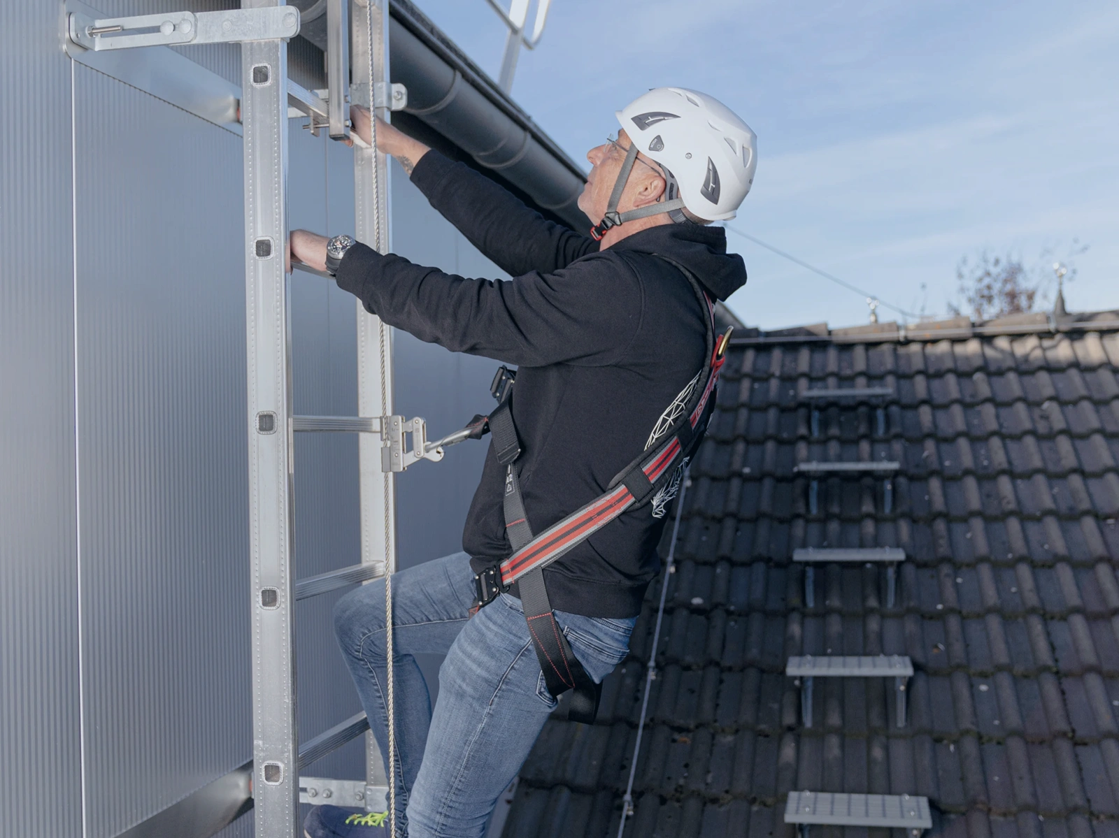 Person med faldsikringsudstyr klatrer på fast stige med vertikalt livlinesystem og glider, som sikrer kontinuerlig faldsikring under op- og nedstigning på tag.